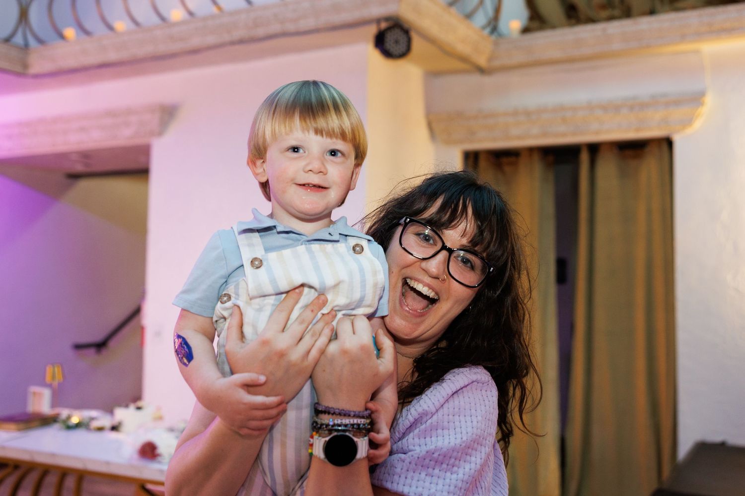 Dark-haired woman with glasses joyfully holding smiling young blonde toddler boy at wedding reception.