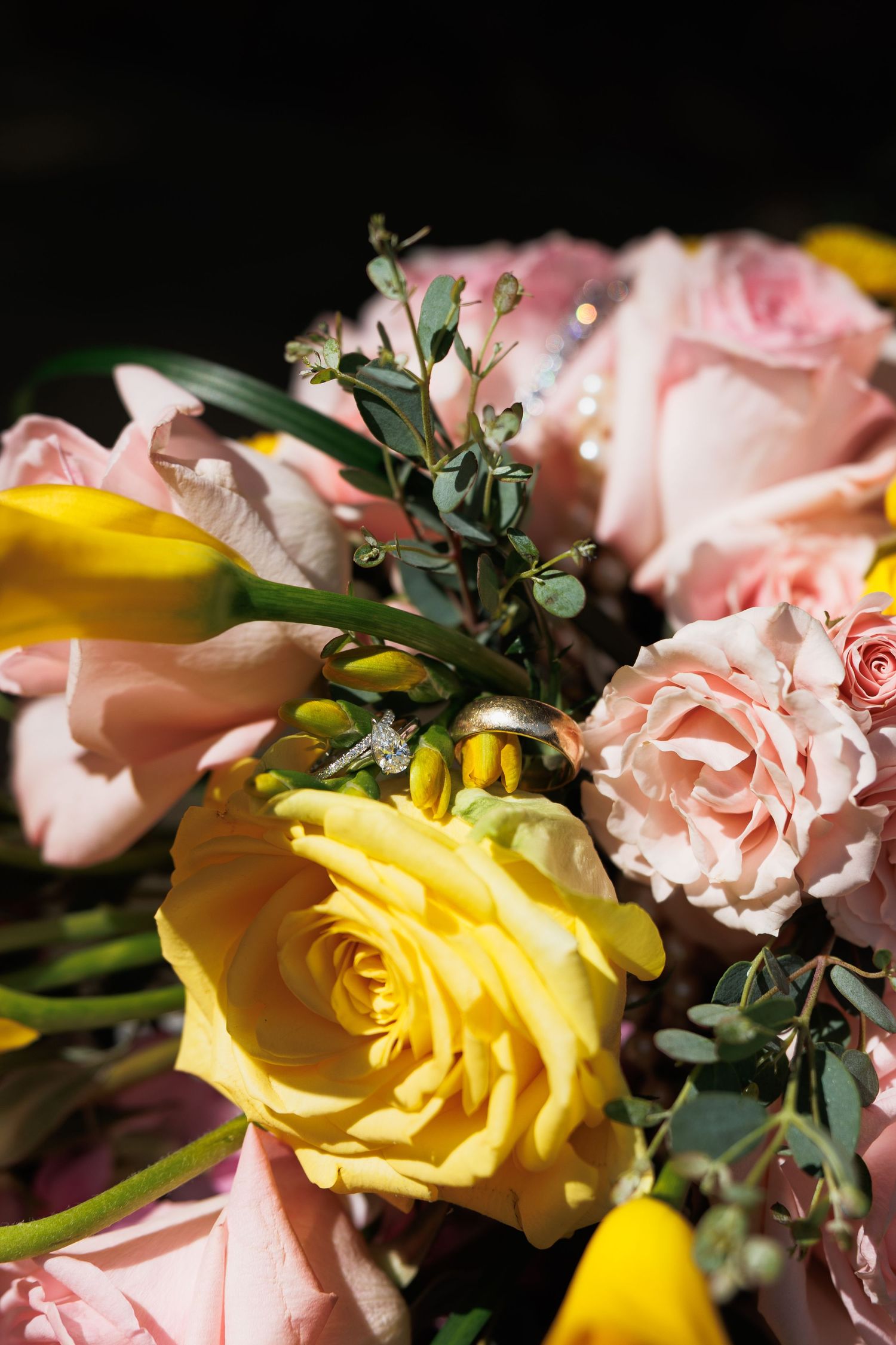 Wedding rings resting on a bouquet of pink and yellow roses with eucalyptus greenery.