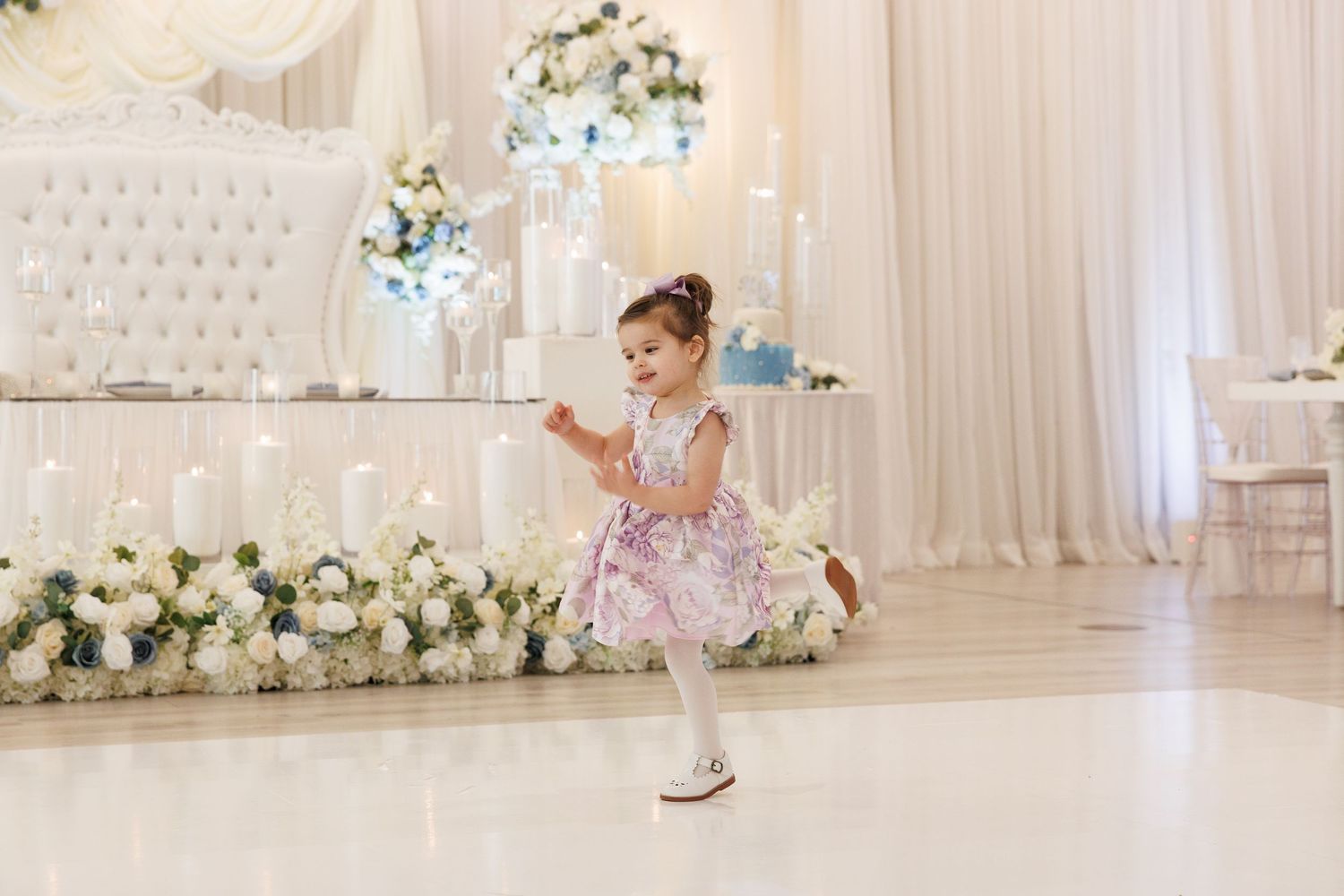 Adorable little girl in pink floral dress dances joyfully on white dance floor at wedding reception.
