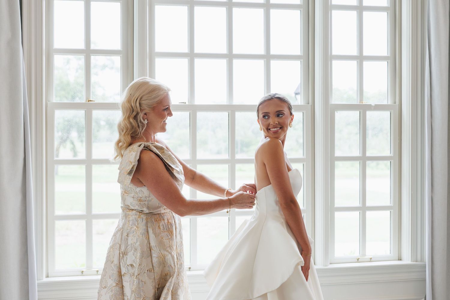 Bridesmaid in floral dress helps bride into her white wedding gown in front of bright sunlit windows.