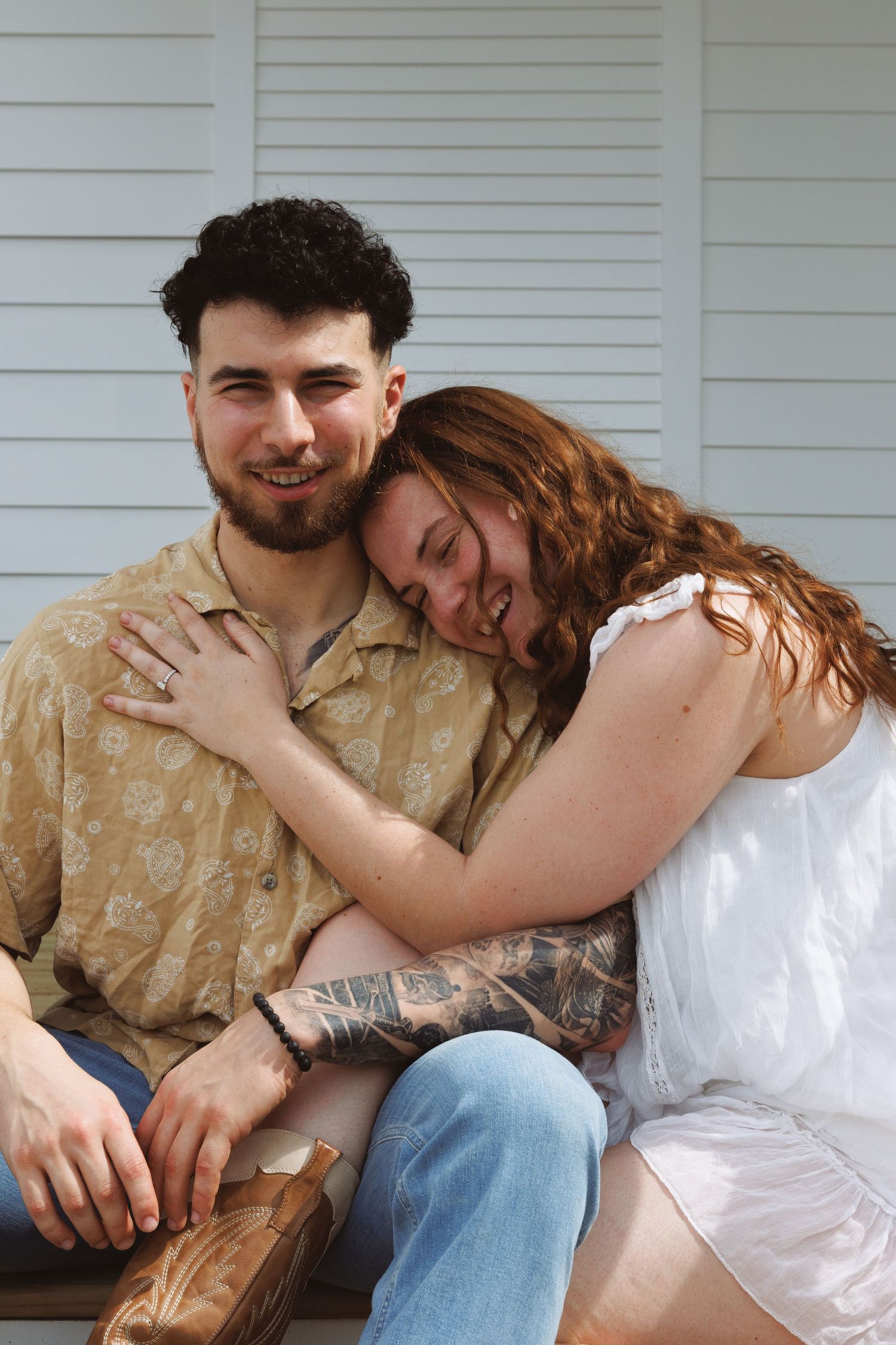 A couple sitting together, woman in white dress leaning on mans shoulder, man in floral shirt smiling, porch background.