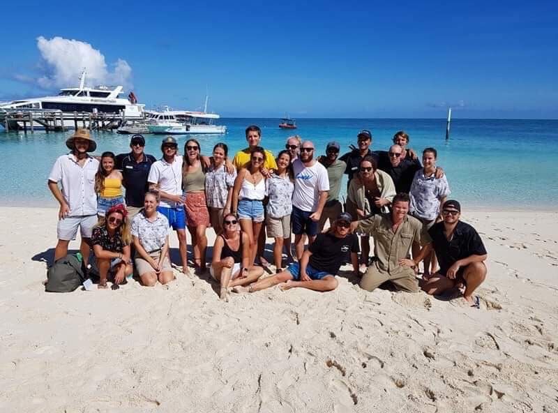 Large group of tourists posing on a white sandy tropical beach with turquoise water and boats in background.