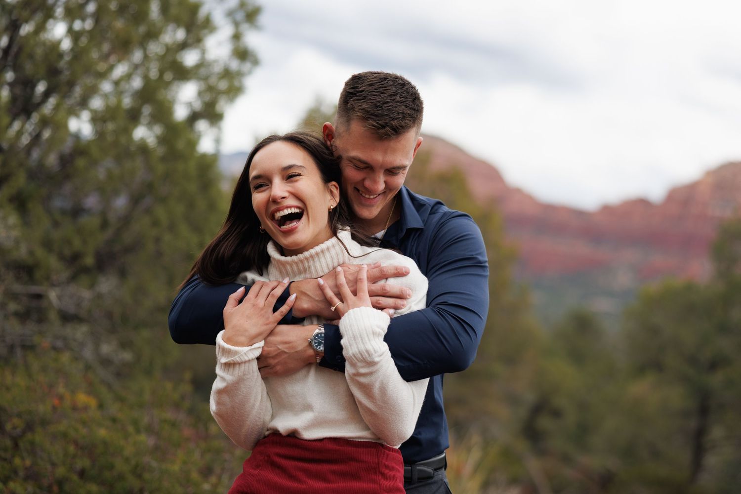Joyful couple embracing outdoors with woman showing engagement ring near red rock formations.