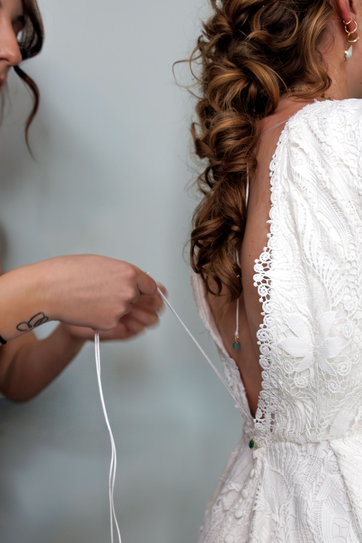 Bridesmaid lacing up the back of a brides white lace wedding gown with curly hair.