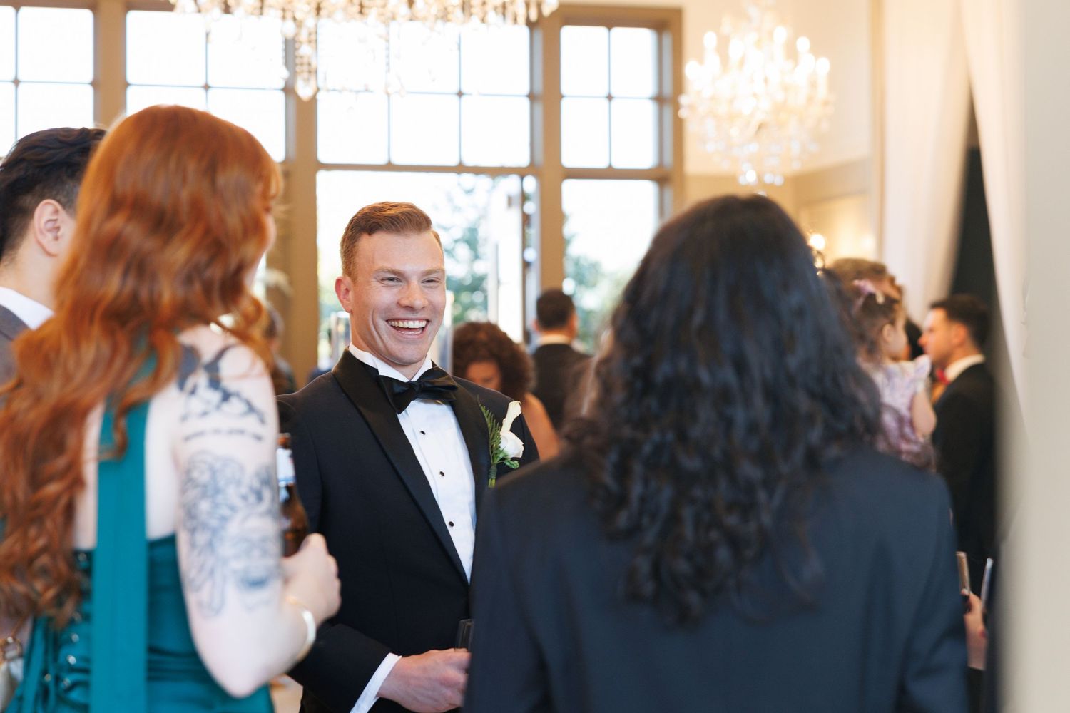 Smiling groom in black tuxedo laughing with wedding guests in elegant chandelier-lit venue.