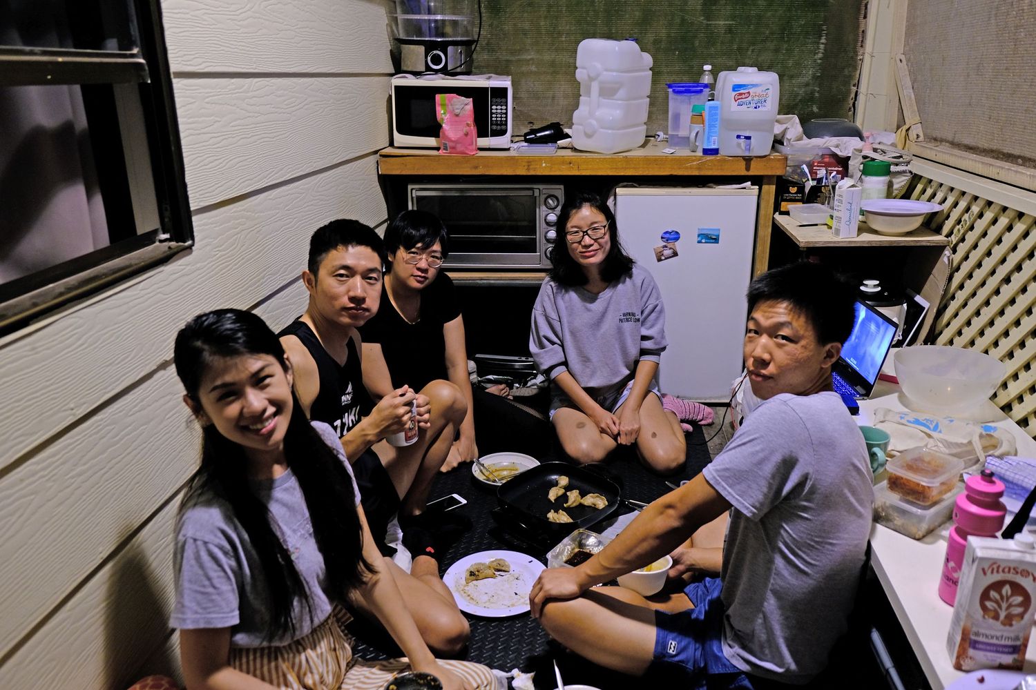 Group of five young Asian friends gathered in a small kitchen, sharing a meal together on the floor.