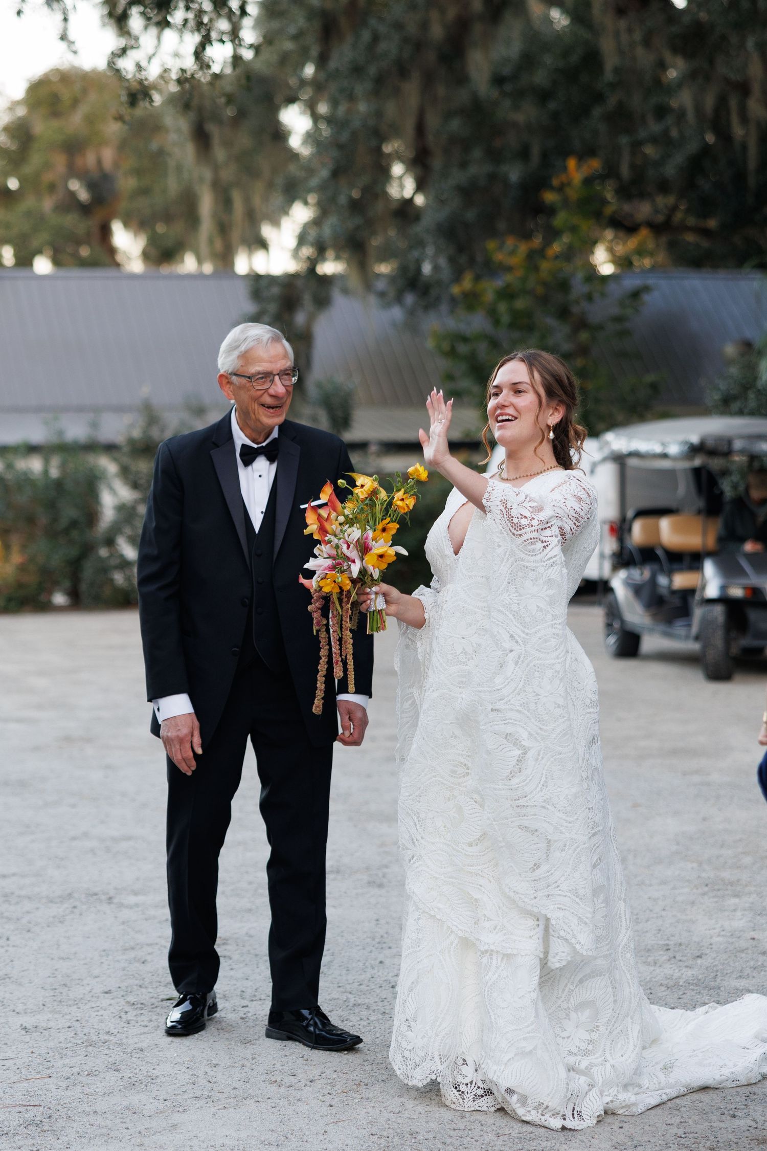 Bride in lace gown holding colorful bouquet waves beside elderly man in tuxedo at outdoor wedding venue.