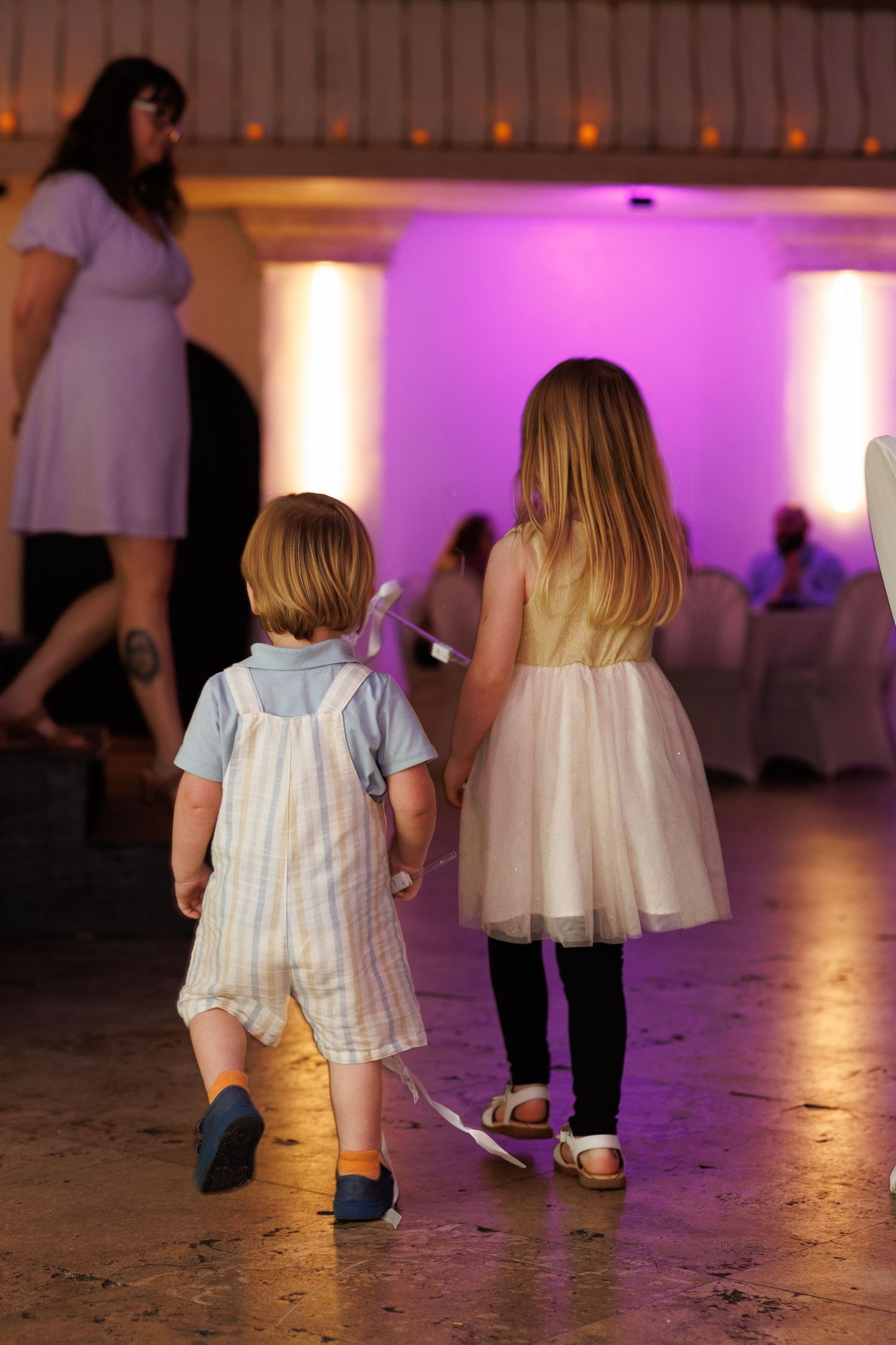 Two young children walking hand in hand on a dance floor at an event with purple ambient lighting.