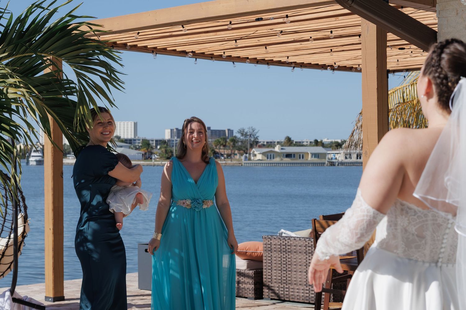 Bride in white gown greeted by woman in teal dress at waterfront wedding venue under wooden pergola.