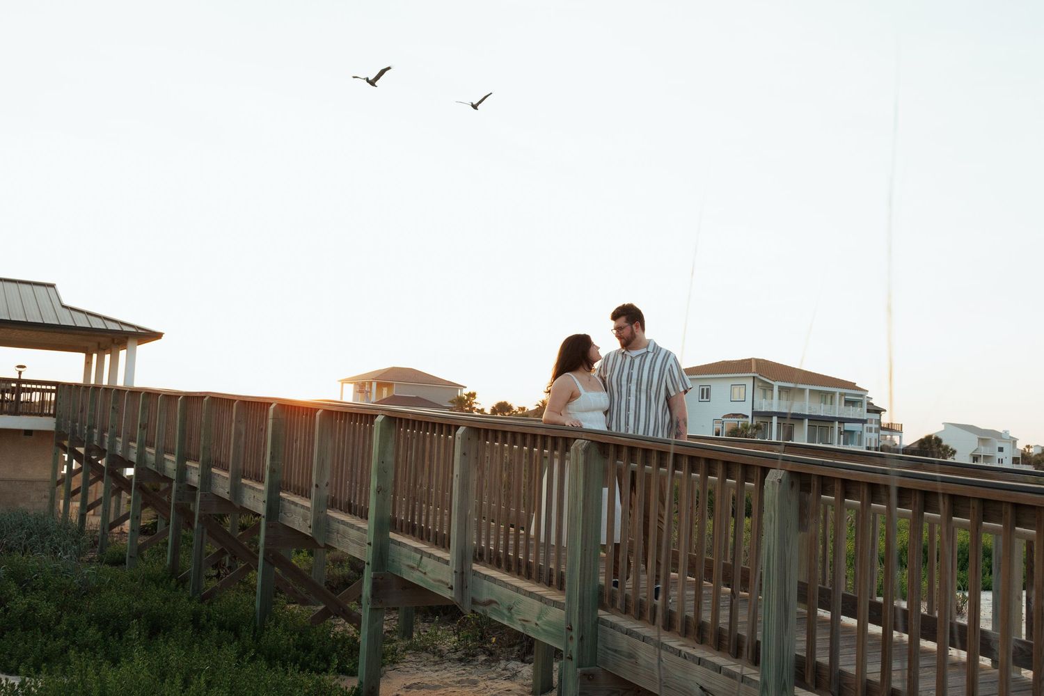Couple standing on a wooden boardwalk near the beach at sunset with birds flying overhead.