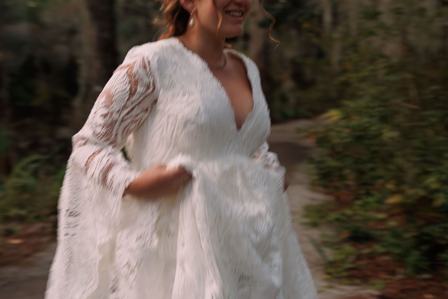 Bride in white lace gown with fur wrap runs joyfully through a wooded forest path.
