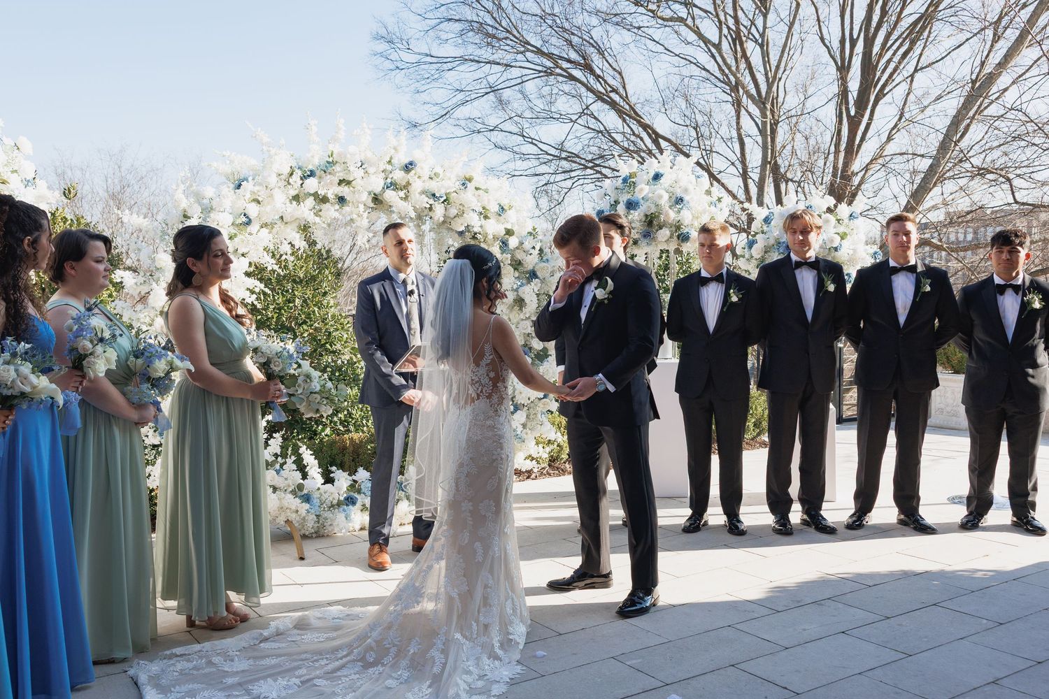 Bride and groom exchange vows at outdoor wedding ceremony with floral arch and wedding party.