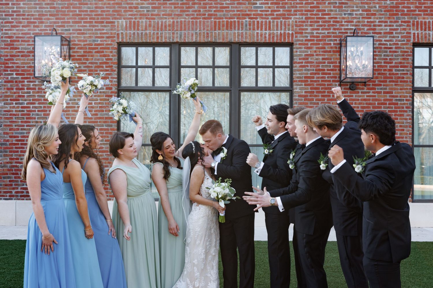 Joyful wedding party celebrates outside brick building, bridesmaids in blue and sage gowns cheering with groomsmen in black tuxedos.