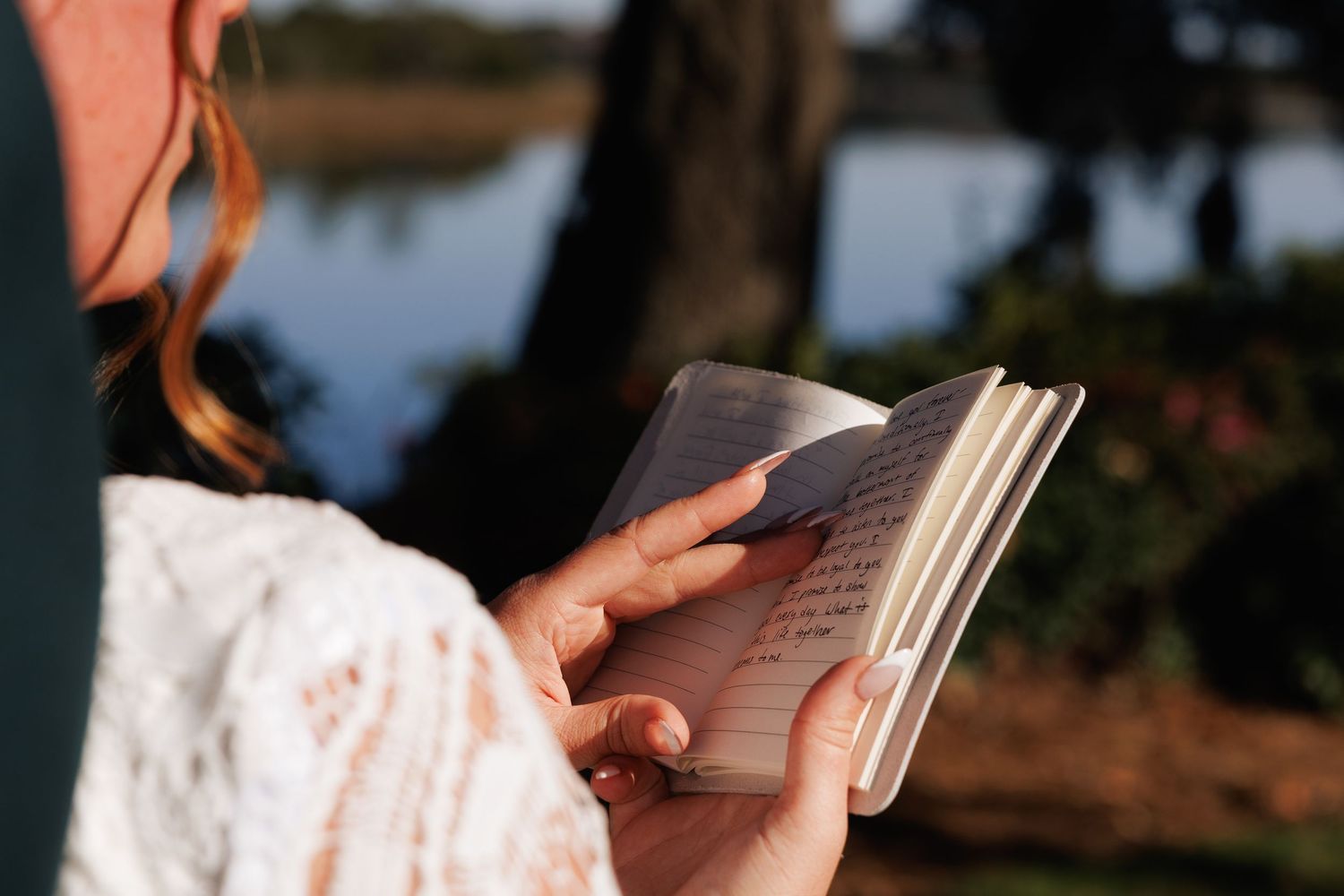 Bride in lace dress reading handwritten vows from a small notebook outdoors by a lake.