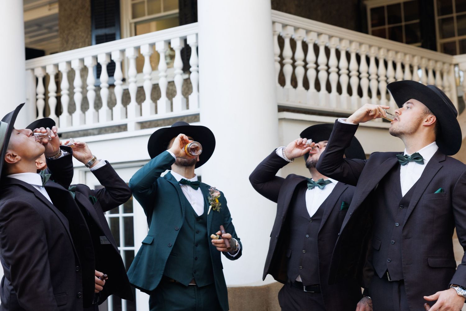 Groomsmen in dark suits and wide-brim hats taking celebratory shots outside a white columned mansion.