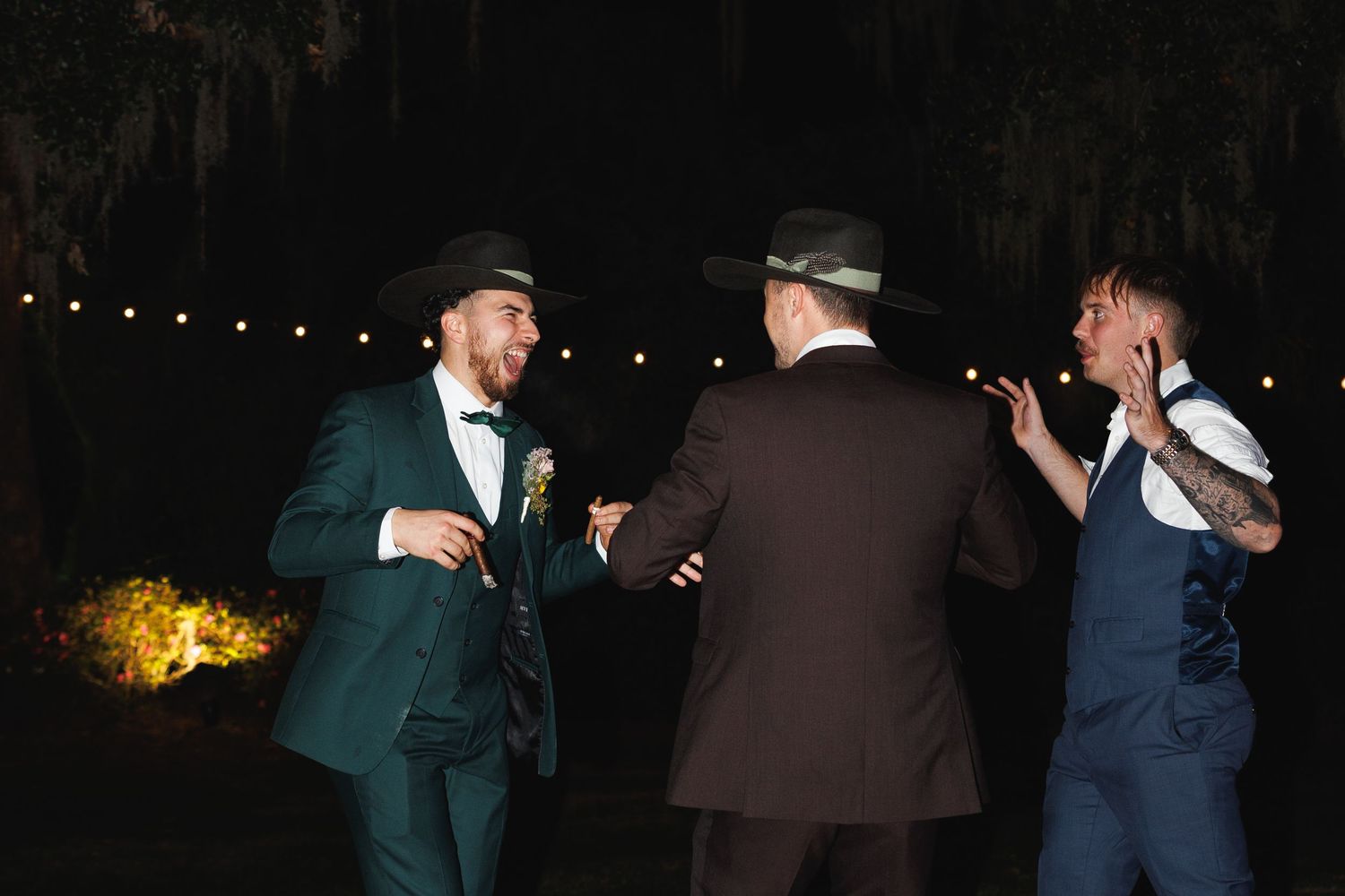 Two grooms in cowboy hats celebrate joyfully with a friend under Spanish moss at night.