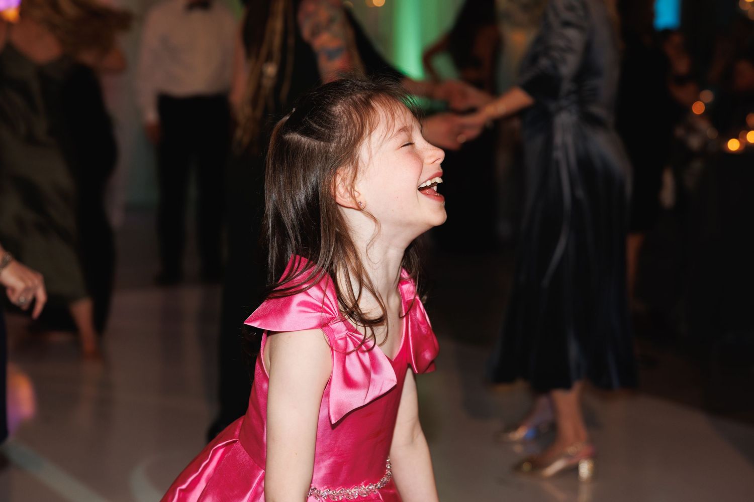 A young girl in a pink satin dress laughs with pure joy while dancing at a wedding reception.