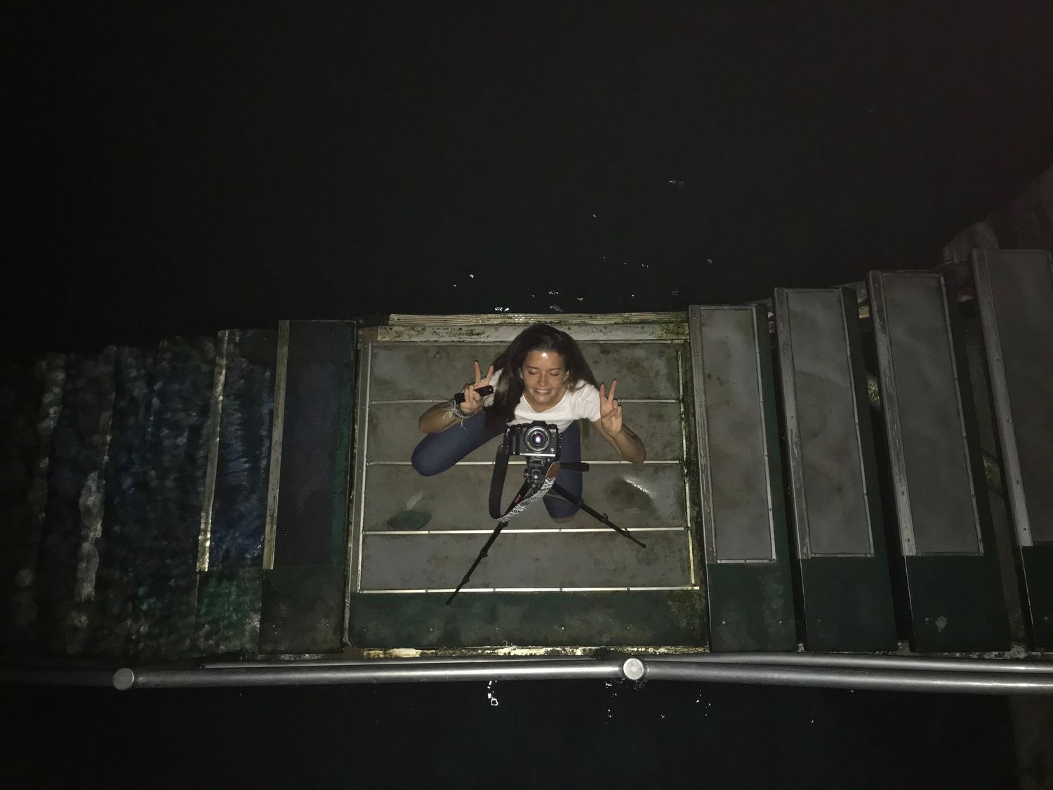 Person making peace sign gesture through a metal gate frame at night outdoors.