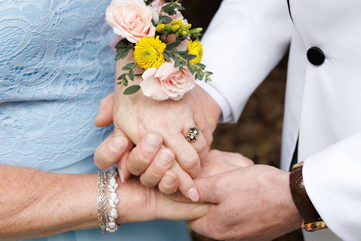 Close-up of hands holding at wedding, woman wearing floral corsage with pink and yellow flowers.