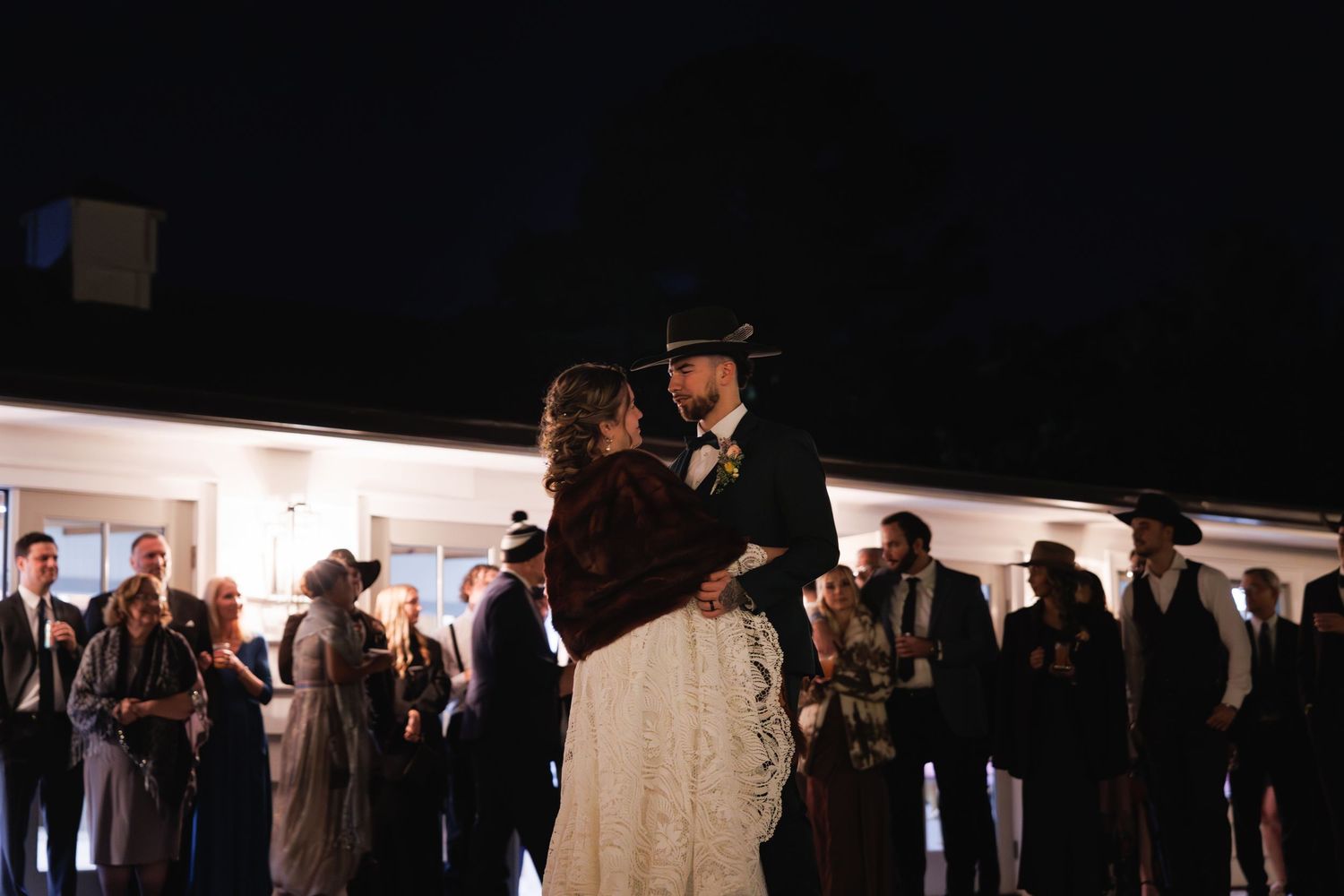 Bride and groom share first dance outdoors at night, surrounded by wedding guests, groom in black cowboy hat.