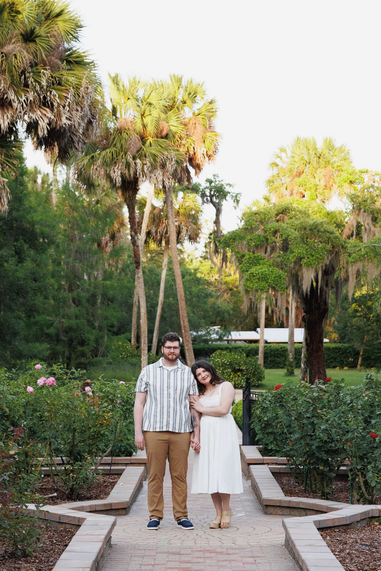 Couple posing together on a garden path with palm trees and roses in background.