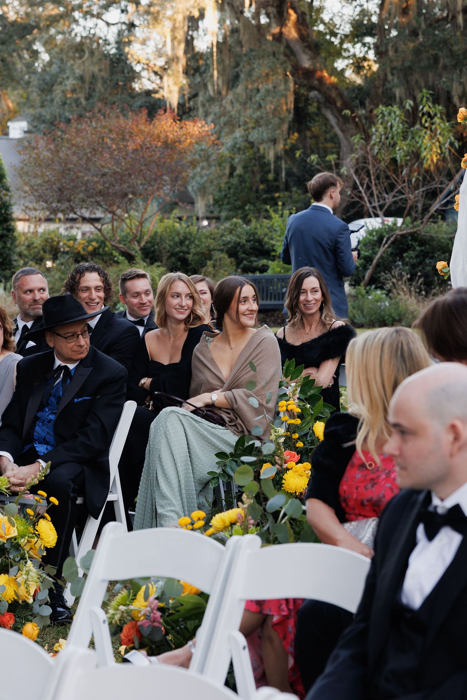 Wedding guests seated in white chairs at an outdoor garden ceremony with colorful yellow and orange floral arrangements.