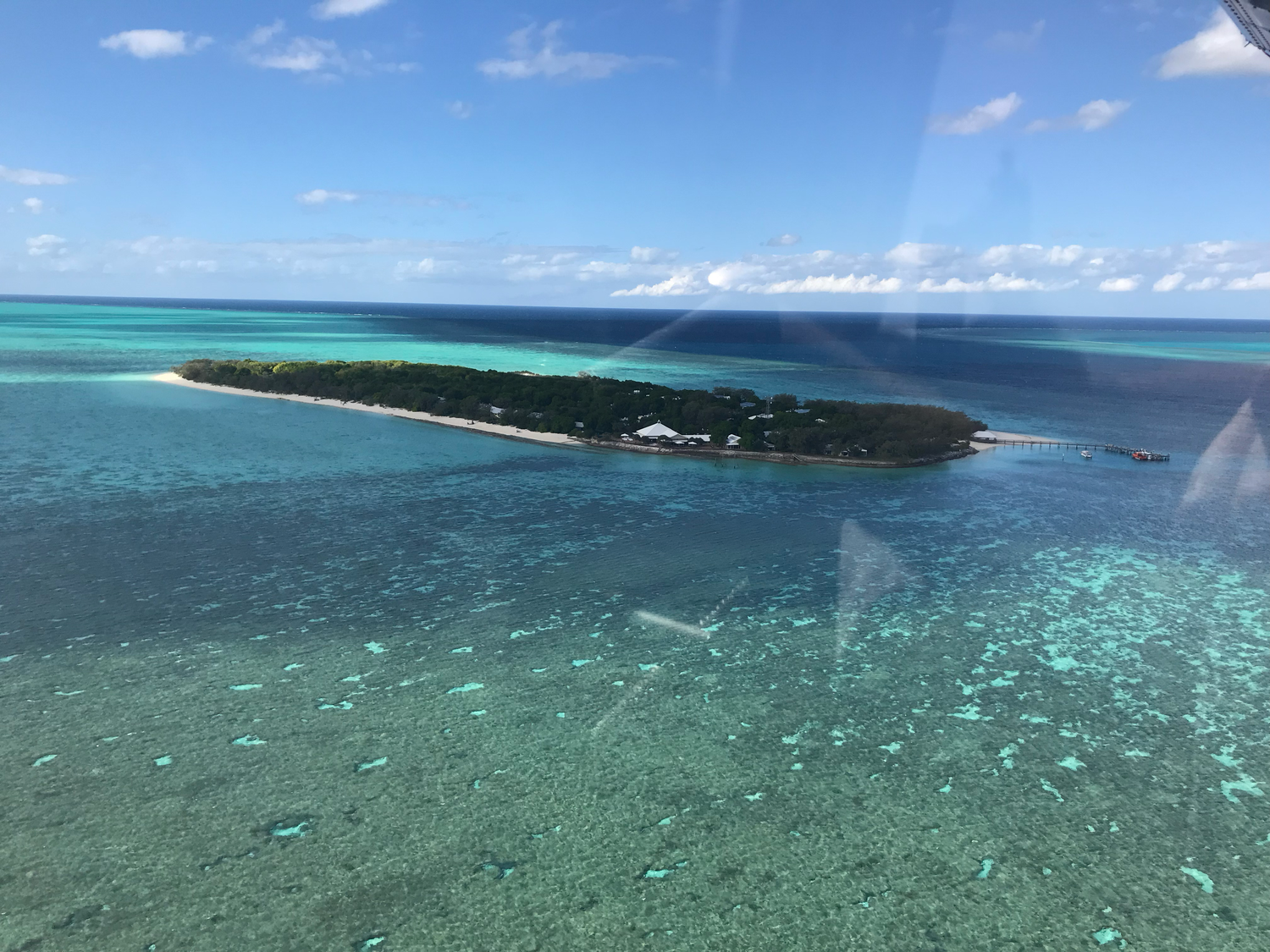 Aerial view of a small tropical island surrounded by vibrant turquoise coral reef waters under a blue sky.