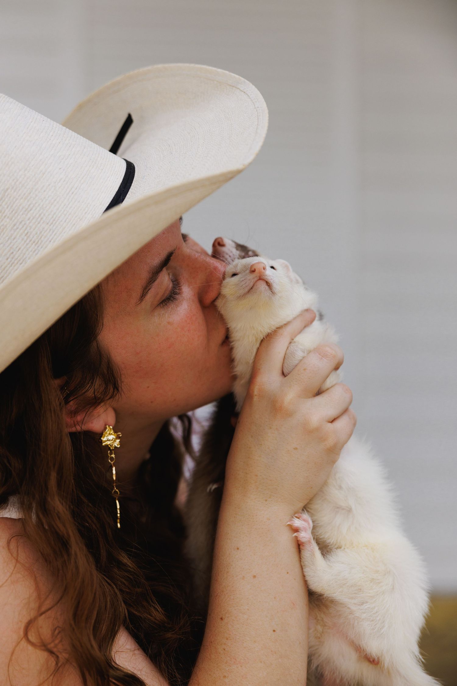Woman in wide-brim hat kissing a white ferret held in her hands.