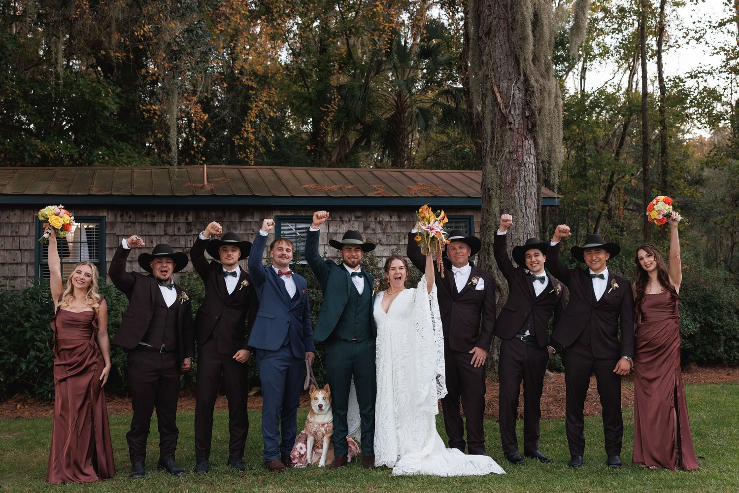 A wedding party of ten people cheers with raised fists outdoors, with a dog sitting at the center of the group.