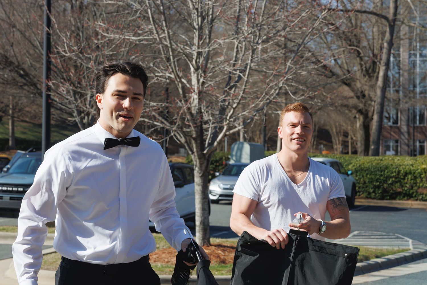 Two well-dressed men walking outdoors near trees, one wearing a bow tie and white shirt, the other in a white t-shirt.