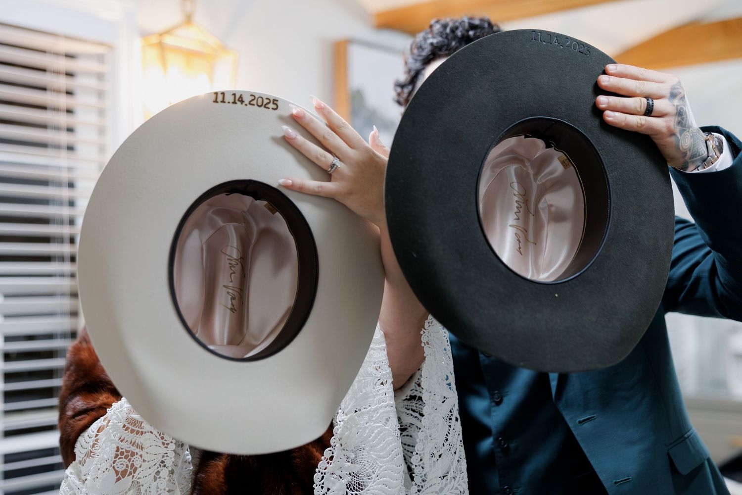 Bride and groom hiding faces behind white and black cowboy hats with wedding date inscribed.