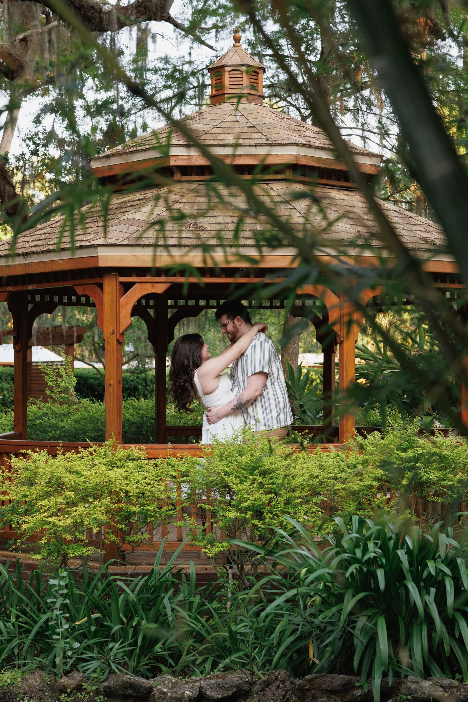Couple embracing romantically inside a wooden gazebo surrounded by lush green gardens.