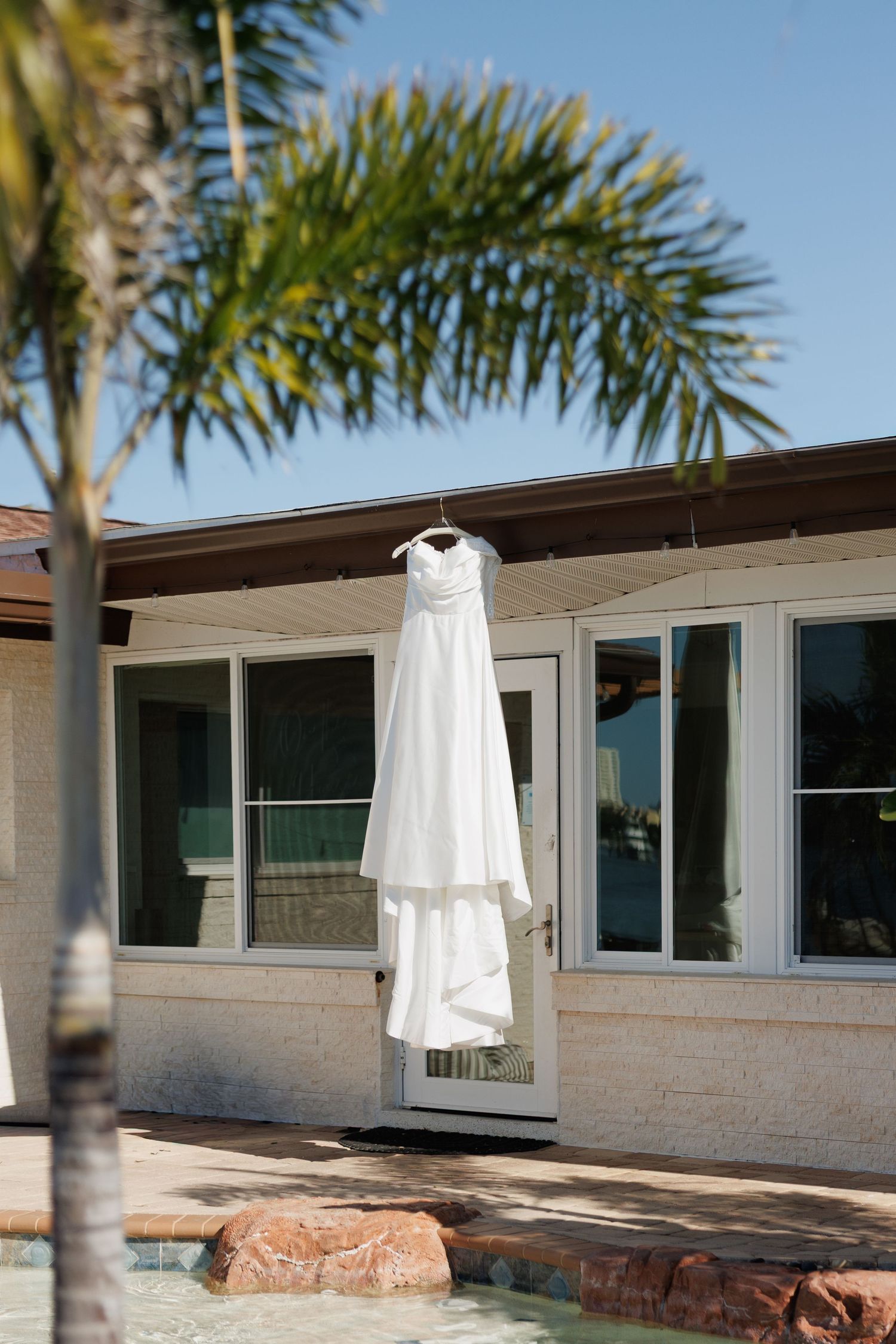 White wedding dress hanging on hanger outside poolside home with palm trees and blue sky.