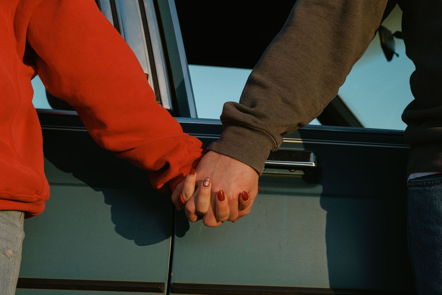 Close-up of couple holding hands in red and gray sweaters beside a blue vehicle outdoors.