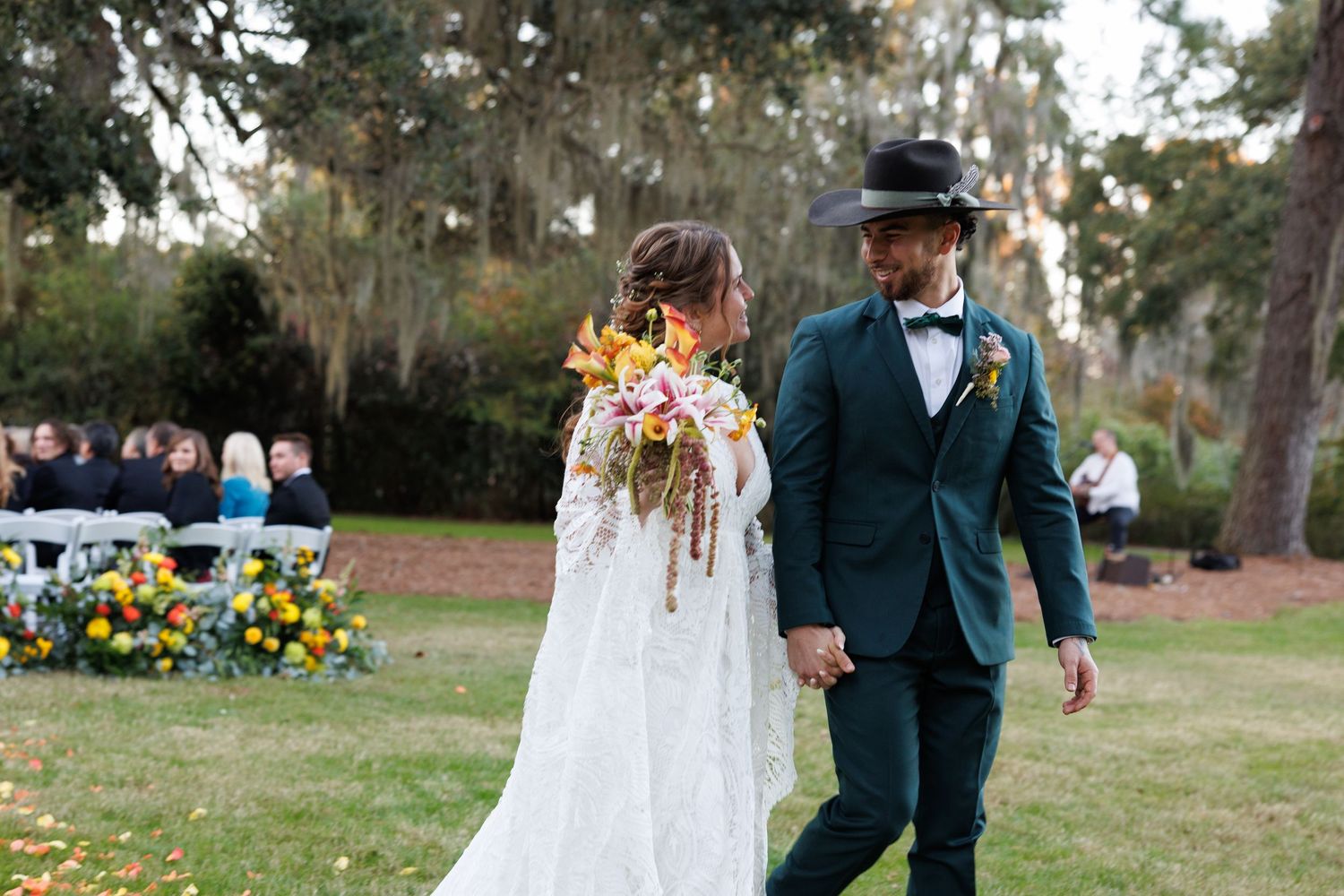 A bride and groom walk hand in hand after their outdoor wedding ceremony, bride holding a colorful floral bouquet.