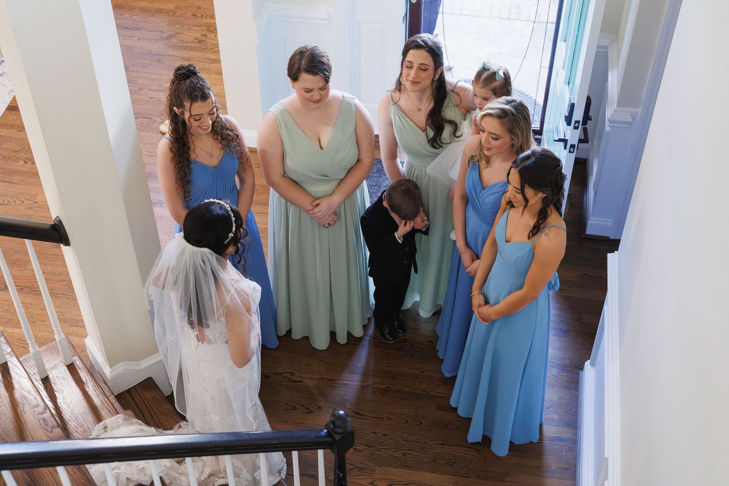 Bridesmaids in blue and sage dresses react to bride's reveal on a staircase.