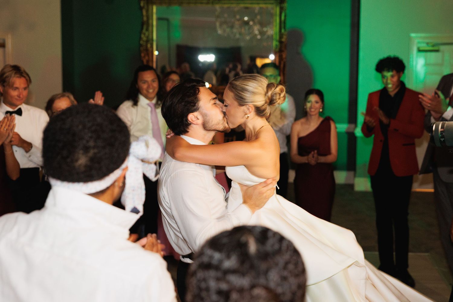 Groom lifts and kisses bride on dance floor surrounded by applauding wedding guests.