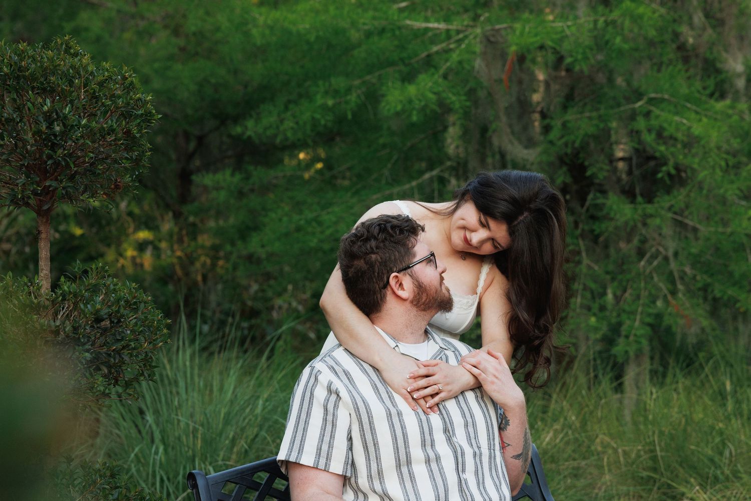 A couple shares a tender moment outdoors, woman embracing man from behind among lush green trees.