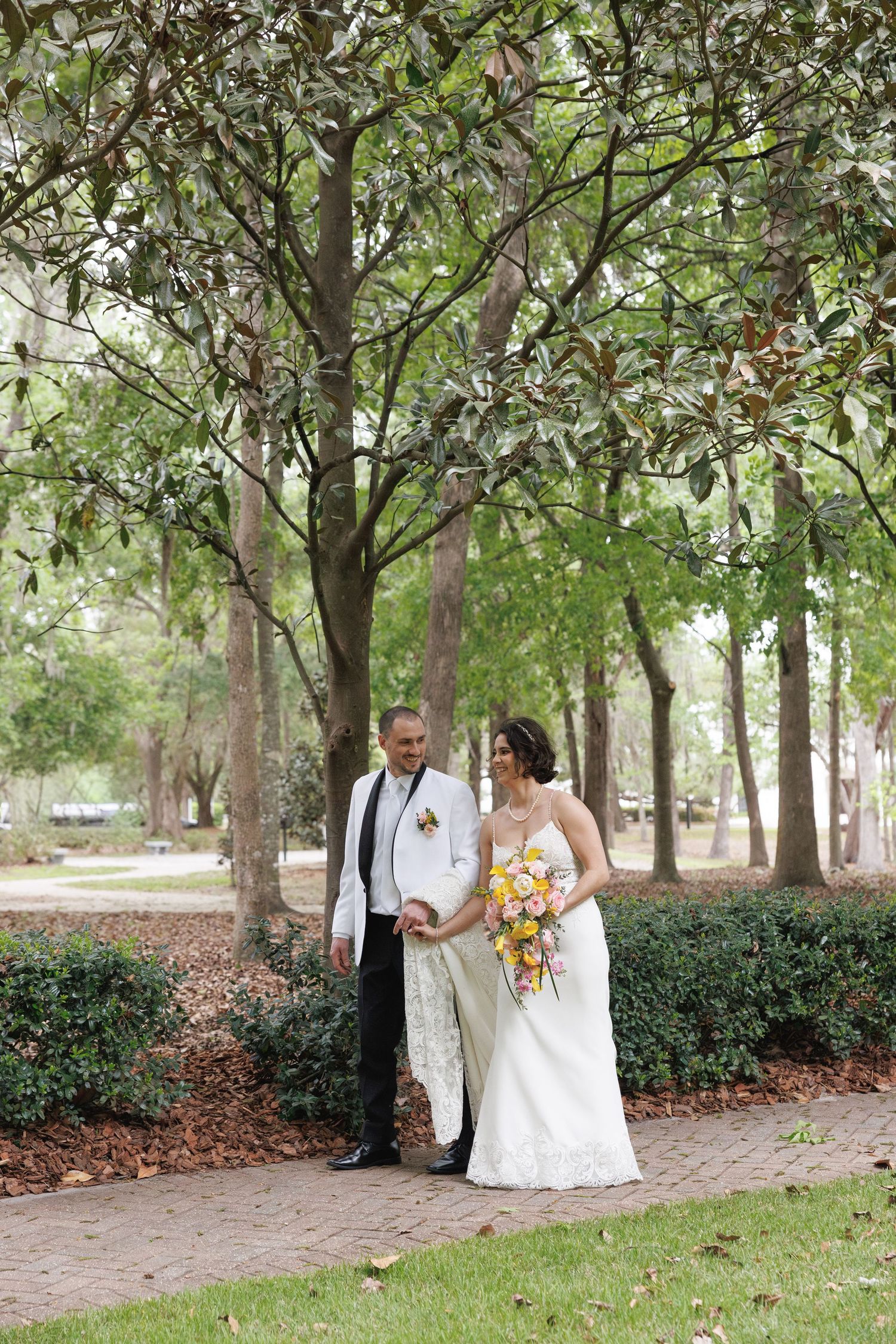 Bride and groom walking together on brick path under canopy of magnolia trees in park.
