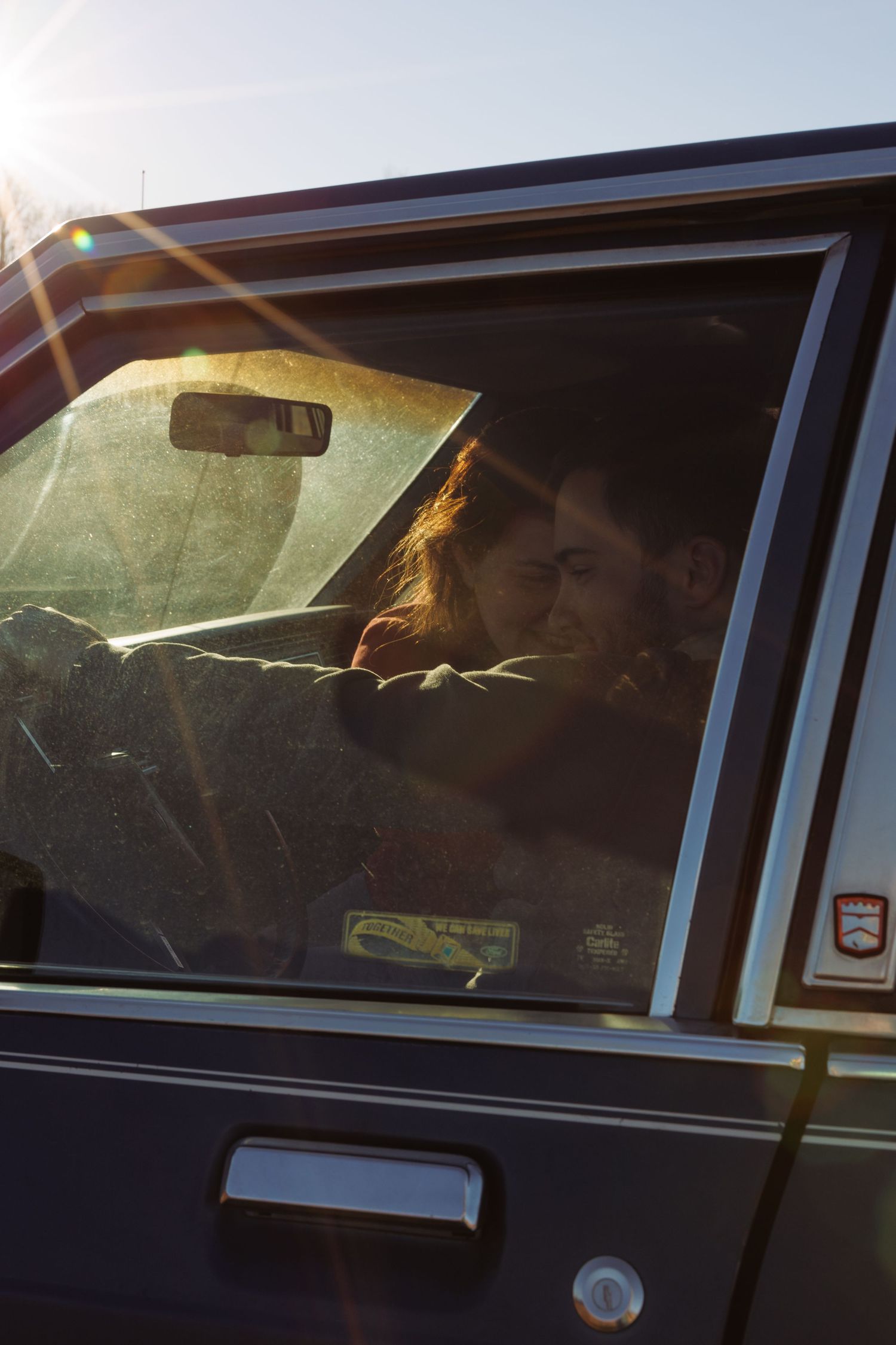 Couple sharing an intimate moment inside a vintage car bathed in golden sunlight with lens flare.