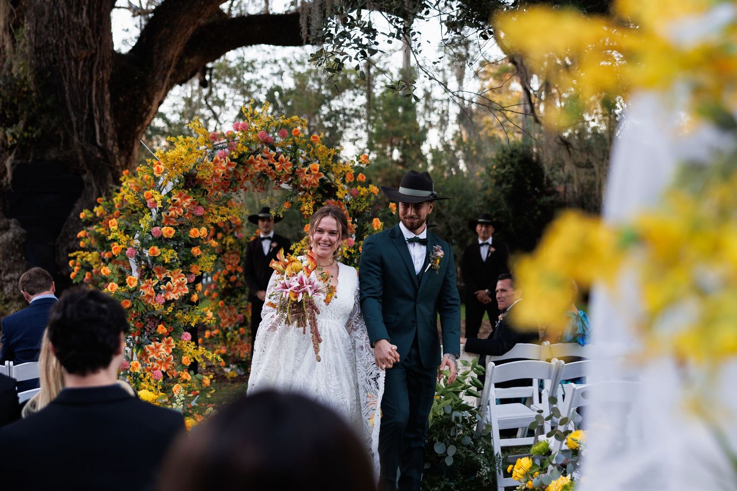 Newlyweds walking down aisle beneath colorful fall floral arch surrounded by wedding guests outdoors.