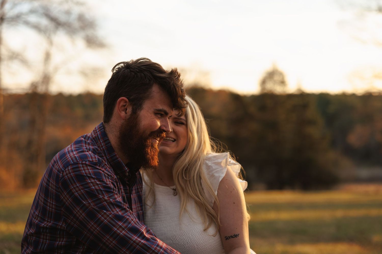 Bearded man and blonde woman share a joyful laugh together during golden hour in an open field.