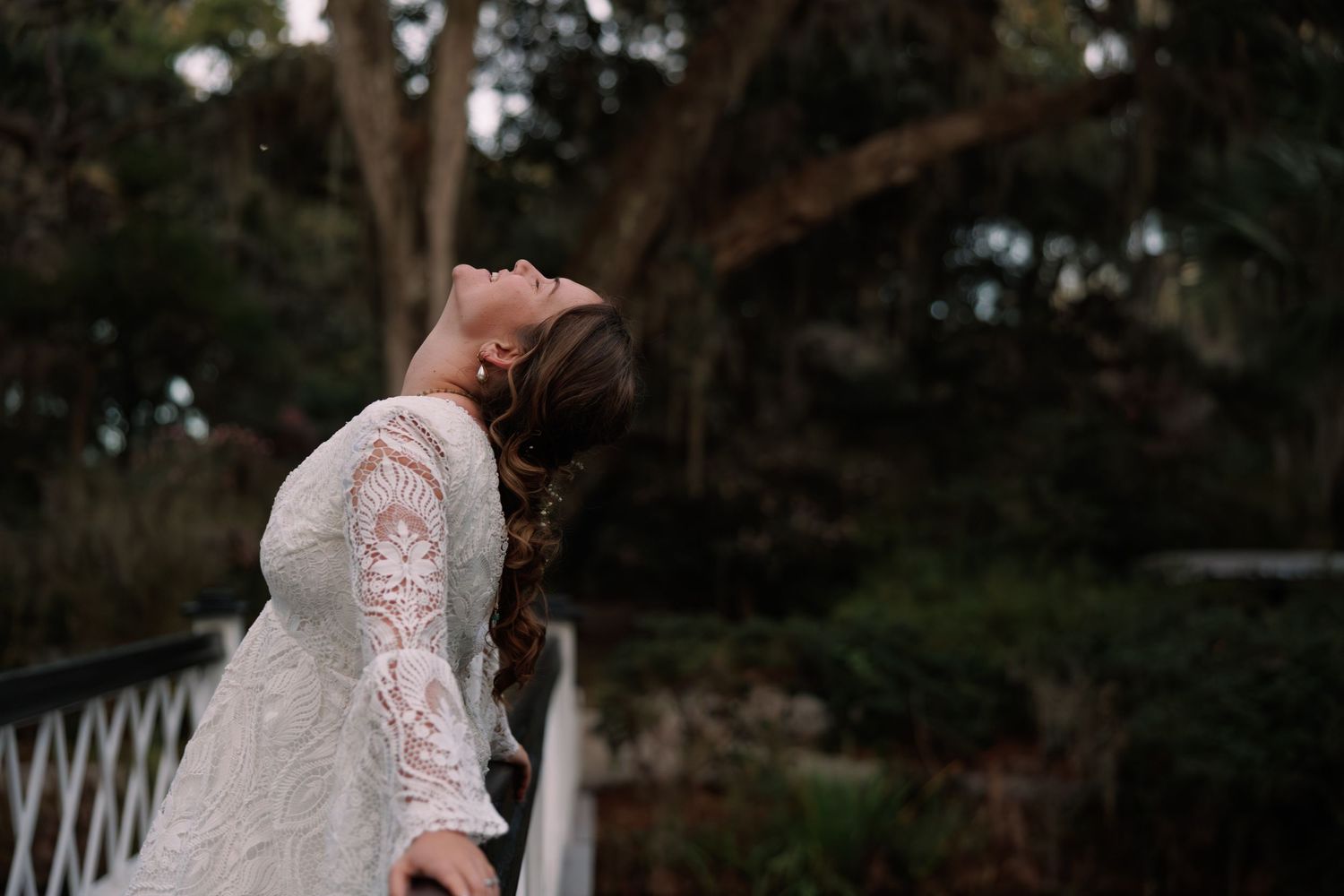 Bride in long-sleeve lace wedding dress tilts head back gracefully on a bridge in a garden.