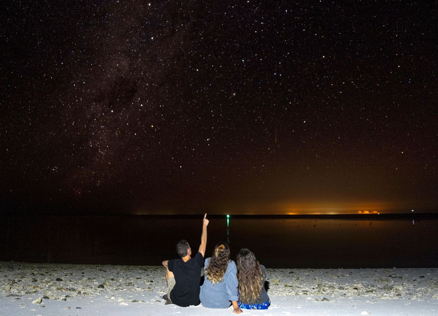 Three friends sit on a beach at night pointing at the Milky Way galaxy in a stunning starry sky.