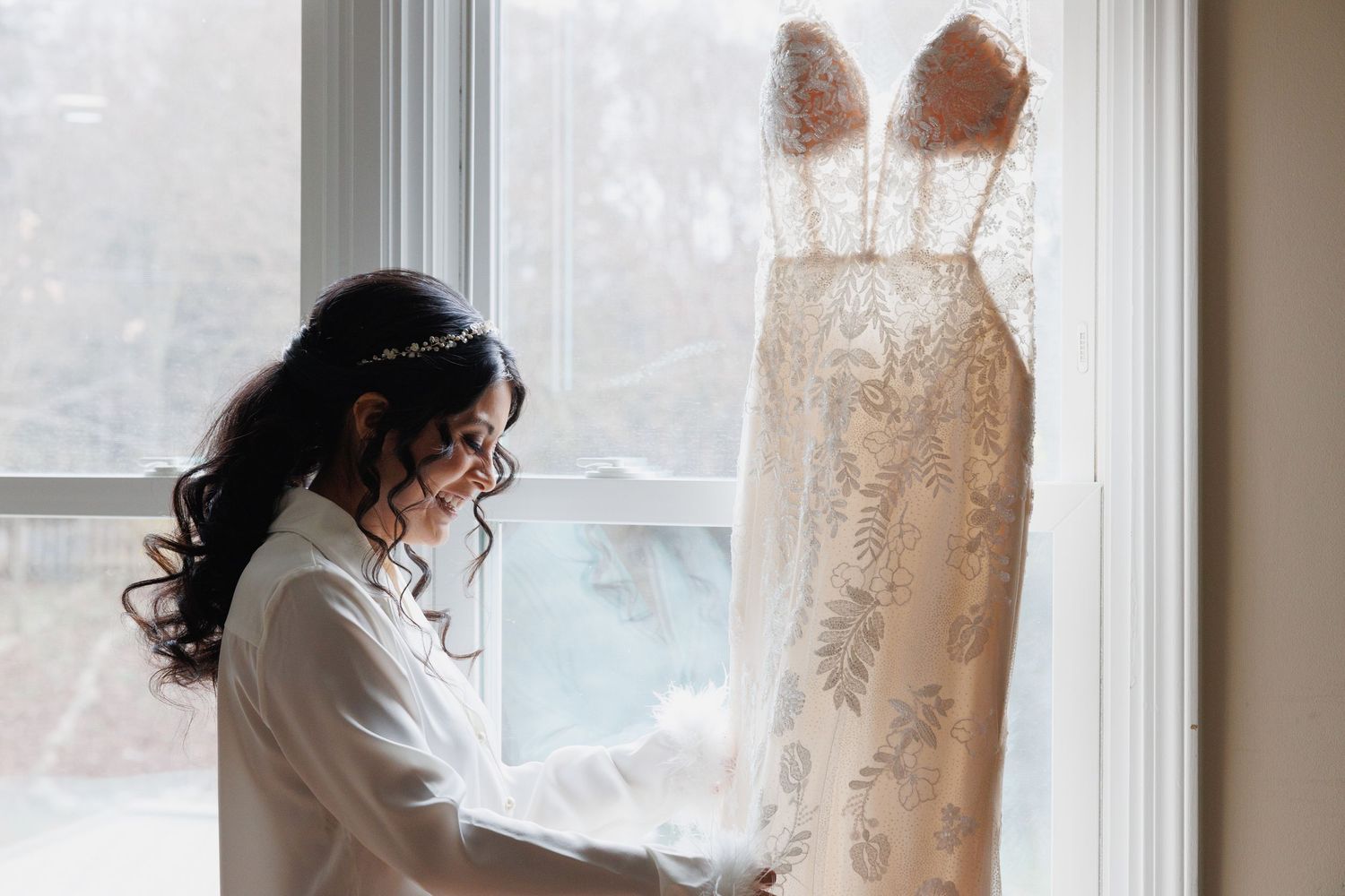 Bride admiring her elegant lace wedding dress hanging by a window in soft natural light.