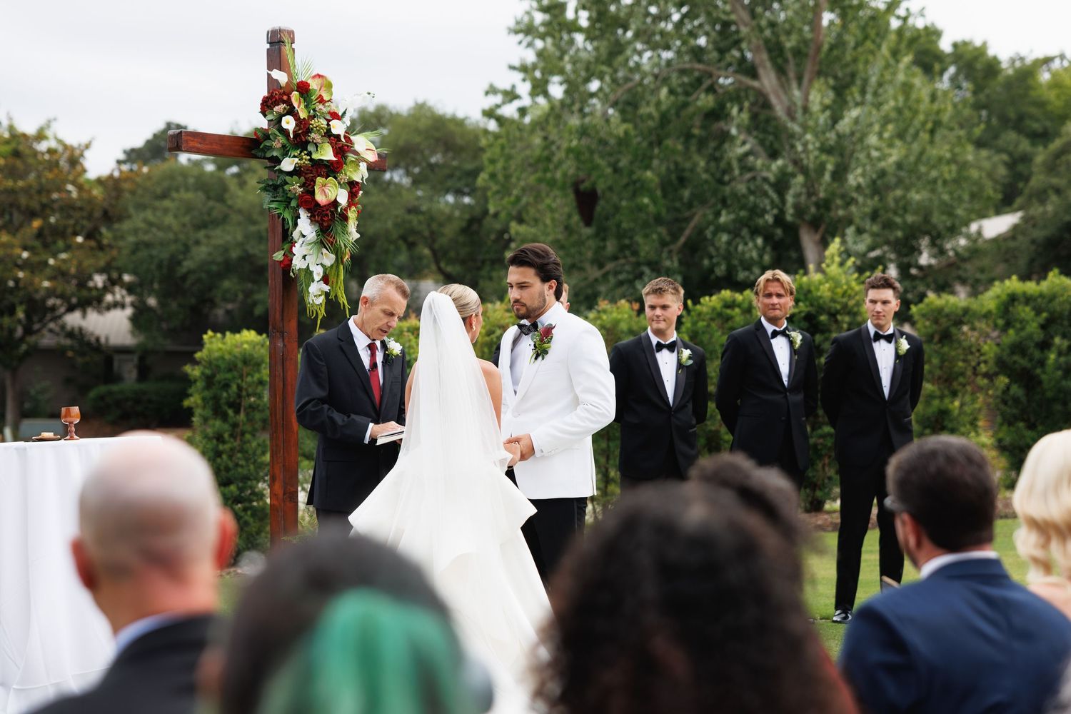Outdoor wedding ceremony with bride and groom at wooden arch adorned with red florals, officiant and groomsmen present.