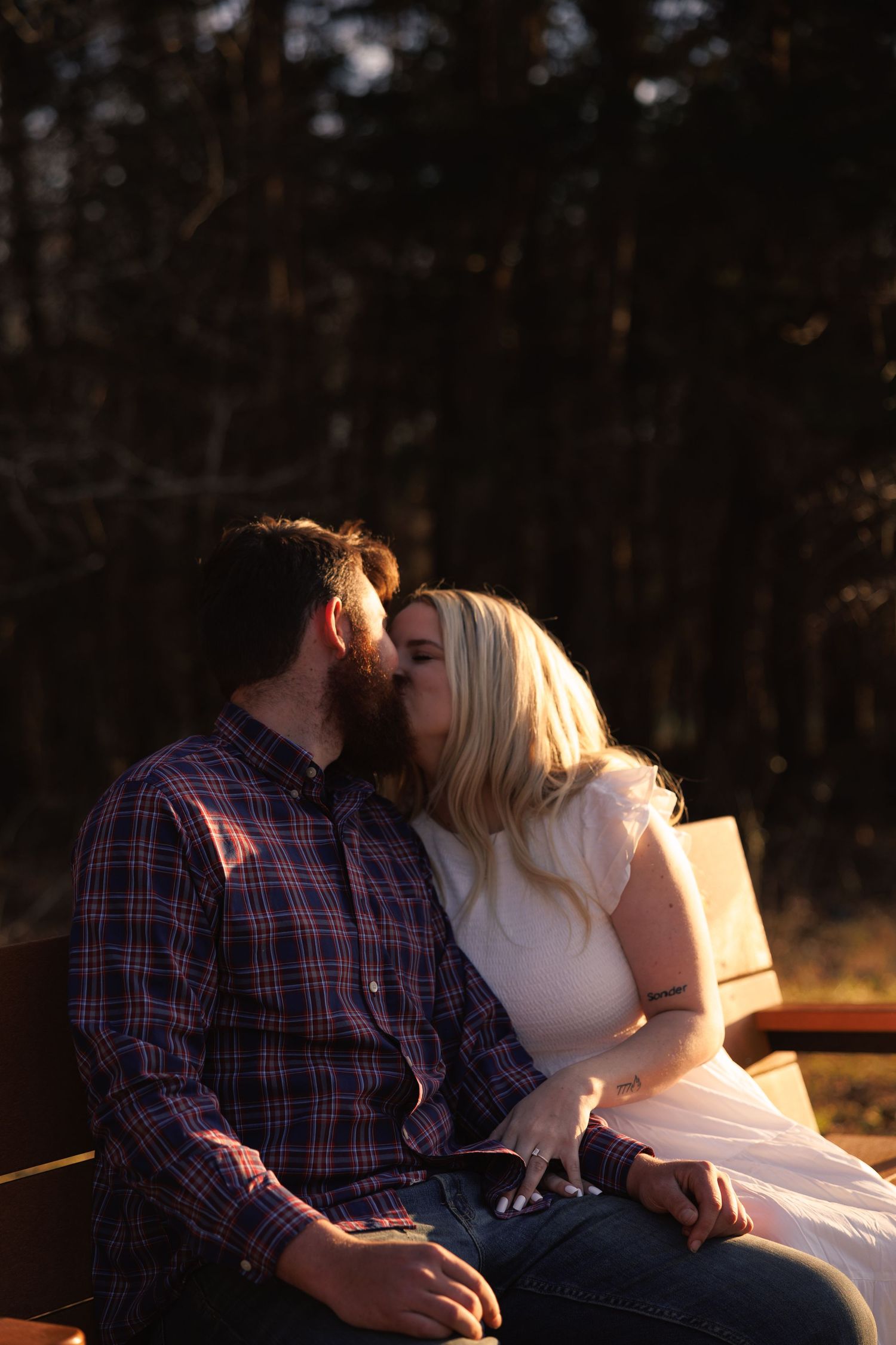Couple sharing a kiss on a wooden bench surrounded by tall trees at sunset.