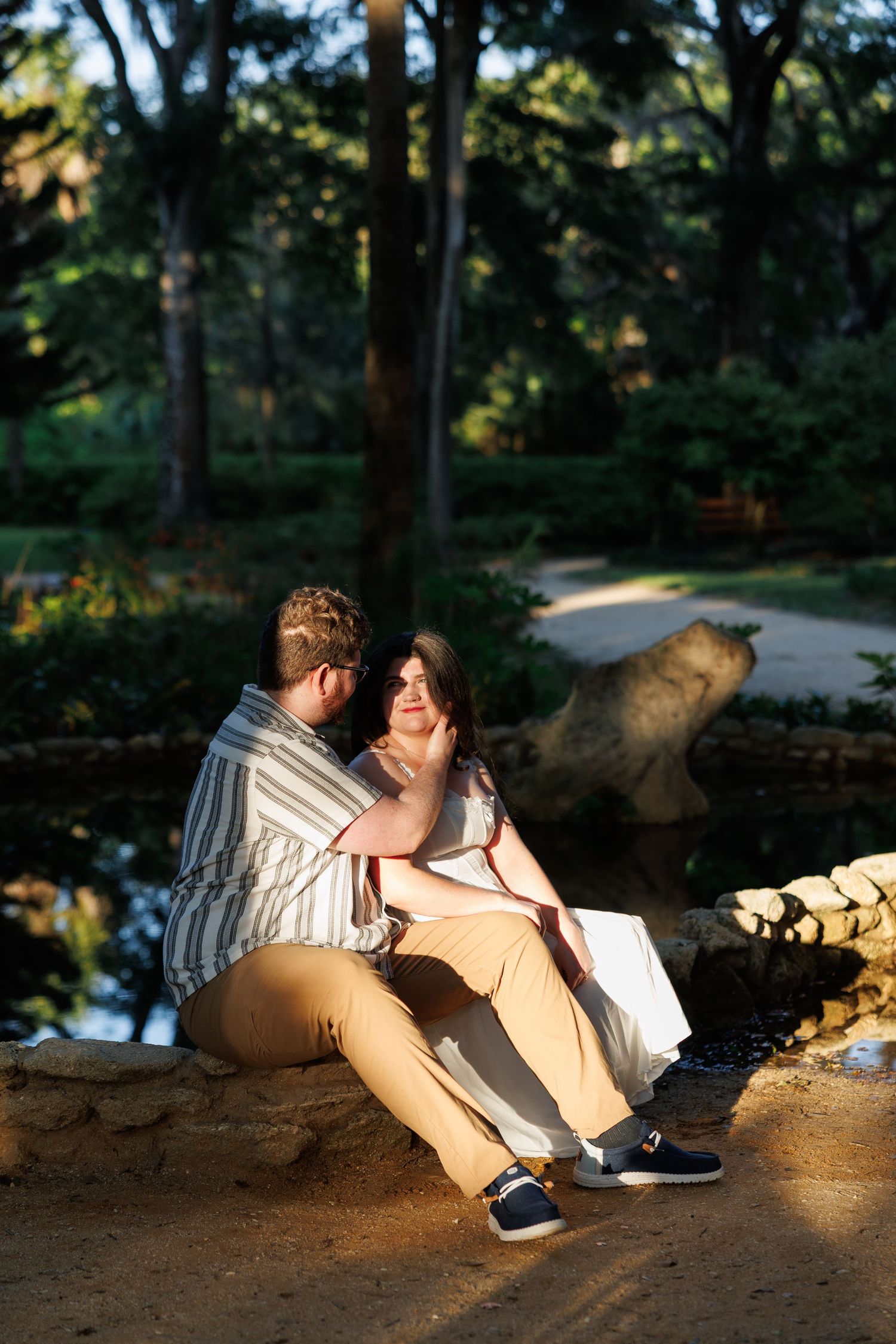 Couple sitting together on stone wall in sunlit park, man tenderly touching womans face in romantic moment.