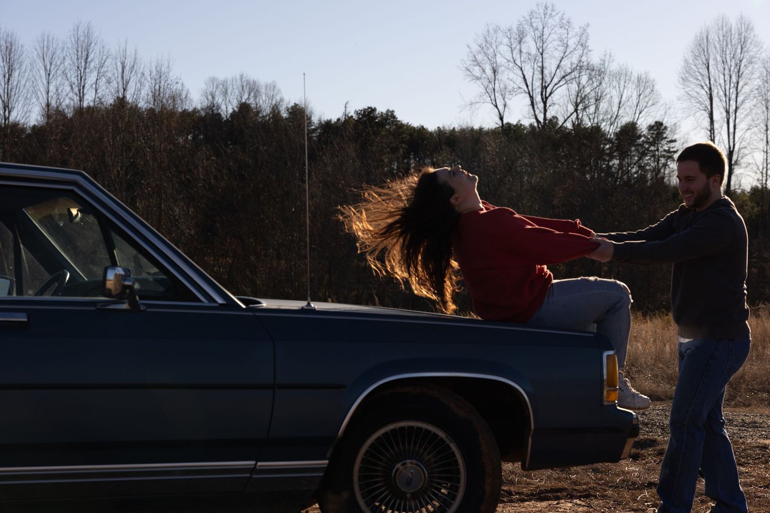 A woman in a red sweater leans back on a vintage blue car while a man stands beside her outdoors.