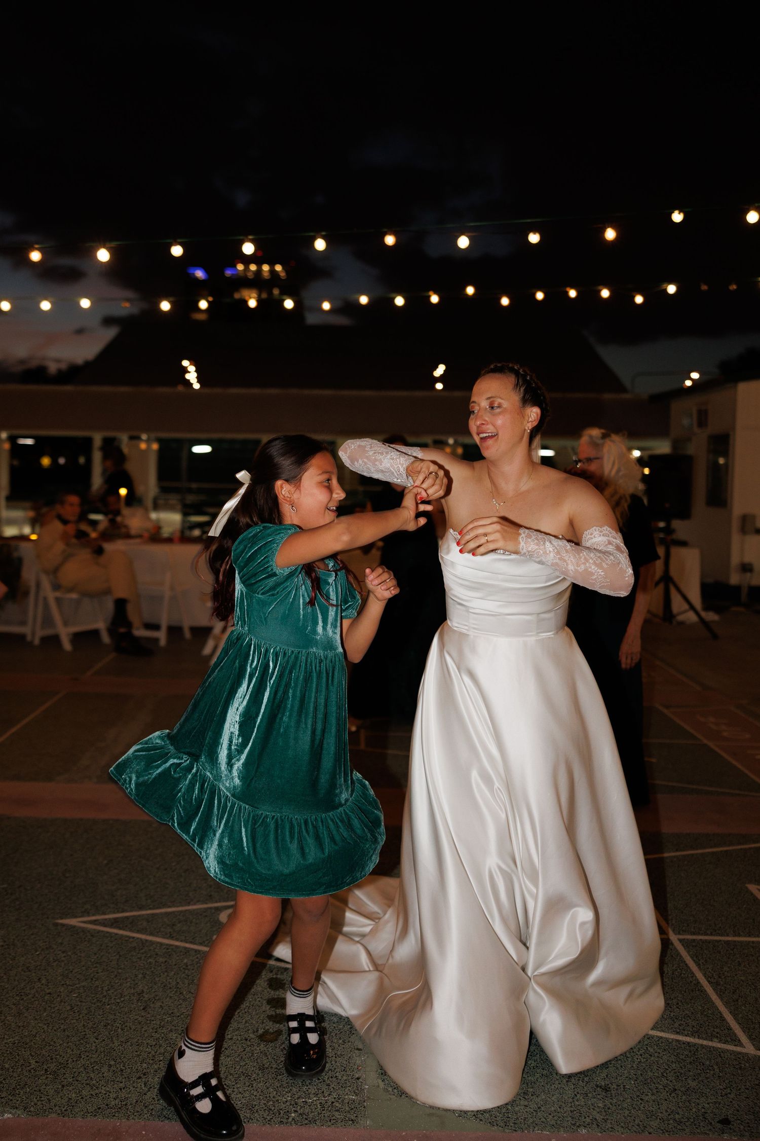 Bride in white satin gown dances joyfully with young girl in green dress at night reception.
